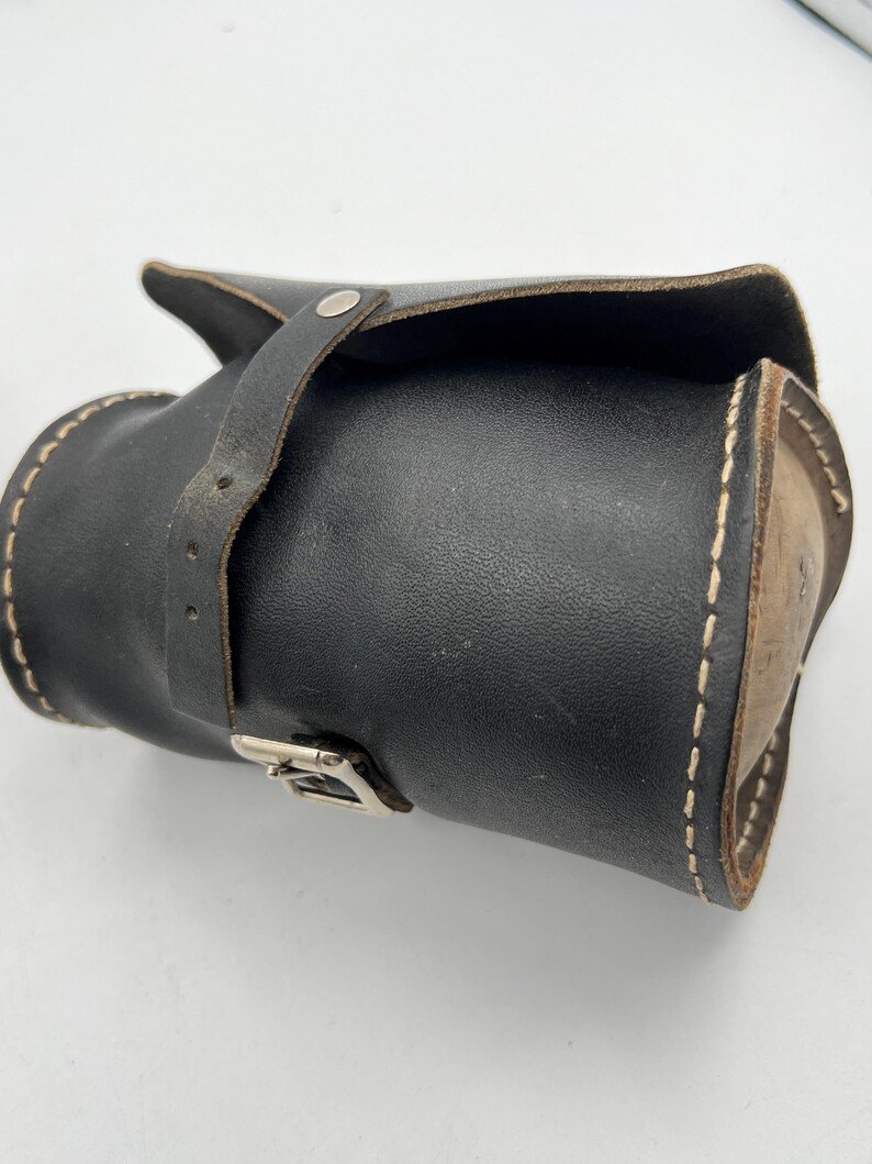 Pode incluir: Um estojo de couro preto com aba e fecho de correia. O estojo cil&iacute;ndrico apresenta costuras brancas e uma fivela prateada. &Eacute; projetado para guardar ferramentas ou objetos pequenos. Aproximadamente 20 cm de comprimento.
