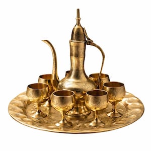 May include: A gold-colored tea set with a decorative floral pattern. The set includes a tall, ornate teapot with a curved spout and handle, six small goblets, and a round tray. The tea set is made of metal and has a shiny finish.