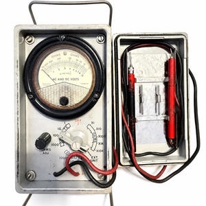 May include: A vintage analog multimeter in a gray metal case with a circular gauge labeled OHMS and AC AND DC VOLTS. It features a central white rotary dial, a black OHMS ADJ knob, and a side compartment containing two red and black test leads.