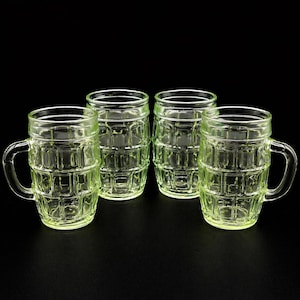 May include: Four clear, green-tinted glass beer mugs with a barrel-like design. Each mug has a handle and a thick, sturdy base. The mugs are arranged on a black surface, creating a simple, yet elegant presentation.