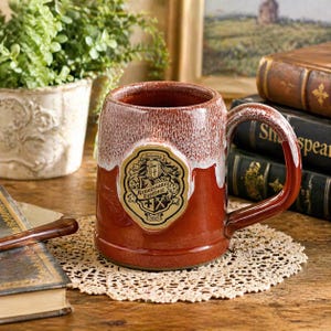 May include: A brown ceramic mug with a large handle, featuring a white and brown glaze. The mug has a gold emblem with the words "Arizona Renaissance Festival" and the year 2015. The mug is sitting on a cream-colored doily.