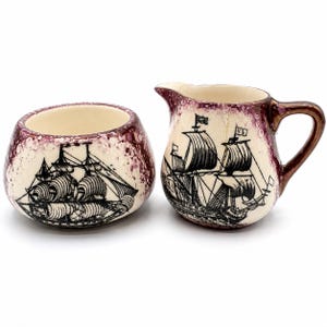 May include: A cream-coloured ceramic sugar bowl and creamer set. Each piece features a black illustration of a sailing ship. The rims are decorated with a speckled maroon pattern. The creamer has a handle and spout.
