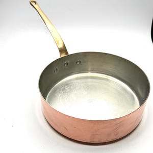 May include: A round copper sauté pan with a polished metallic interior and a long, curved brass handle attached to the side with three visible rivets. The exterior of the pan features a warm copper finish, while the interior is silver-toned.