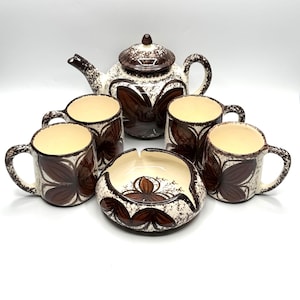 May include: A vintage ceramic tea set featuring a teapot with lid, four mugs, and an ashtray. The set has a cream-colored base with brown floral designs and accents. The mugs have handles and the ashtray is round.
