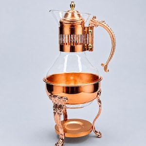 May include: A copper-colored coffee maker with a clear glass carafe and lid. The coffee maker has an ornate handle and is supported by decorative legs. The design is vintage and elegant.