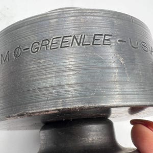 Pode incluir: Close-up de uma ferramenta de metal industrial resistente com o texto "MO-GREENLEE - USA" gravado em sua superf&iacute;cie. A ferramenta tem uma forma cil&iacute;ndrica com uma textura estriada e um acabamento met&aacute;lico escuro. A ferramenta parece ser usada para fins industriais.