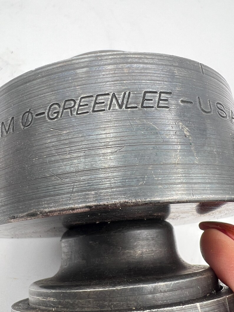 Pode incluir: Close-up de uma ferramenta de metal industrial resistente com o texto "MO-GREENLEE - USA" gravado em sua superf&iacute;cie. A ferramenta tem uma forma cil&iacute;ndrica com uma textura estriada e um acabamento met&aacute;lico escuro. A ferramenta parece ser usada para fins industriais.