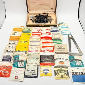 May include: A collection of vintage optical equipment, including a trial frame in a brown case, numerous lens packets, and a metal protractor. The packets display various brand names and lens specifications, with a focus on eye care.