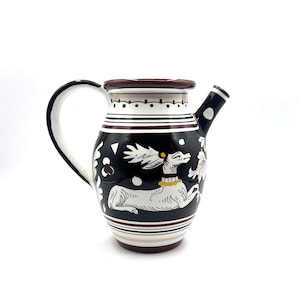 May include: A ceramic pitcher featuring a black background with a white hand-painted deer illustration. The vessel includes a curved handle, a spout, and decorative horizontal stripes in dark red and white around the neck and base.
