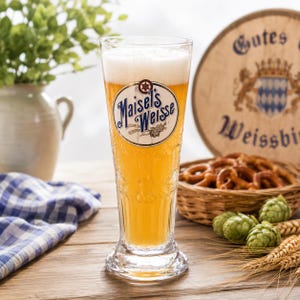 May include: A tall, clear glass filled with golden wheat beer, topped with white foam. The glass features the blue and white Maisel's Weisse logo. The vessel has a textured, embossed surface and a sturdy base, standing upright on a wooden surface.