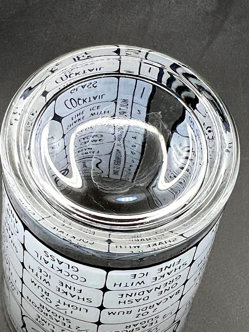 May include: Clear glass cocktail shaker with a white and black label featuring cocktail recipes. The shaker has a wide mouth and a cylindrical body. The text on the label includes cocktail names and ingredient measurements.