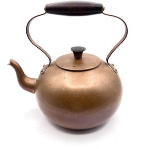 May include: A vintage copper kettle with a dark wooden handle and lid knob. The kettle has a rounded body, a curved spout, and a metal handle that arches over the top. The design is classic and timeless.
