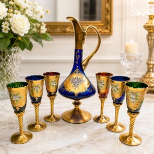 May include: A glass decanter and six matching stemmed glasses featuring deep blue and green glass bodies with gold-painted accents, ornate gold handles, and hand-painted floral motifs in white and green, set on a polished marble surface.