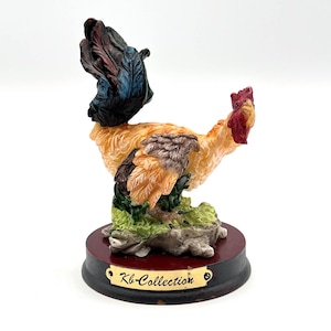 May include: A detailed figurine of a rooster with a red comb, orange and brown feathers, and a black tail. The rooster stands on a base with the text "Kb-Collection". The base is dark red and black.
