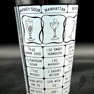May include: A vintage cocktail shaker glass with black and white graphics. The glass features recipes for Whiskey Sour, Manhattan, and Bacardi cocktails, including ingredient measurements in ounces and teaspoons. The glass is clear and cylindrical.