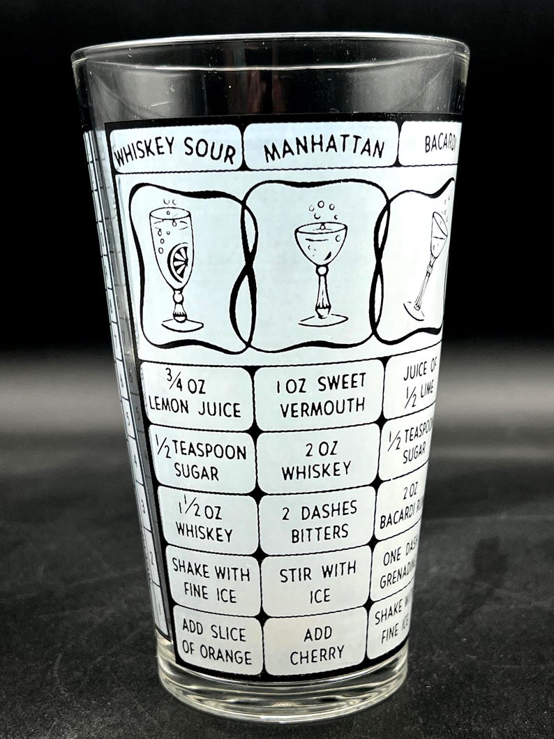 May include: A vintage cocktail shaker glass with black and white graphics. The glass features recipes for Whiskey Sour, Manhattan, and Bacardi cocktails, including ingredient measurements in ounces and teaspoons. The glass is clear and cylindrical.
