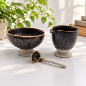May include: A ceramic set consisting of a wide bowl, a pouring pitcher with a spout, and a small ladle. Each piece features a dark, mottled brown and black glaze on the upper portion and a contrasting matte off-white base and handle.