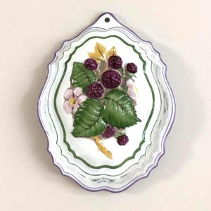May include: White ceramic wall plate with a scalloped edge, featuring a raised design of blackberries, green leaves, and pink flowers. The plate has a green and purple border and a hole for hanging.