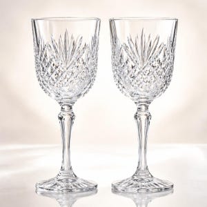 May include: Two clear crystal wine glasses. Each glass features a detailed, cut-glass pattern on the bowl and stem. The bowl has a fan-like design at the top, with diamond-shaped cuts throughout. The stems are slender and elegant.