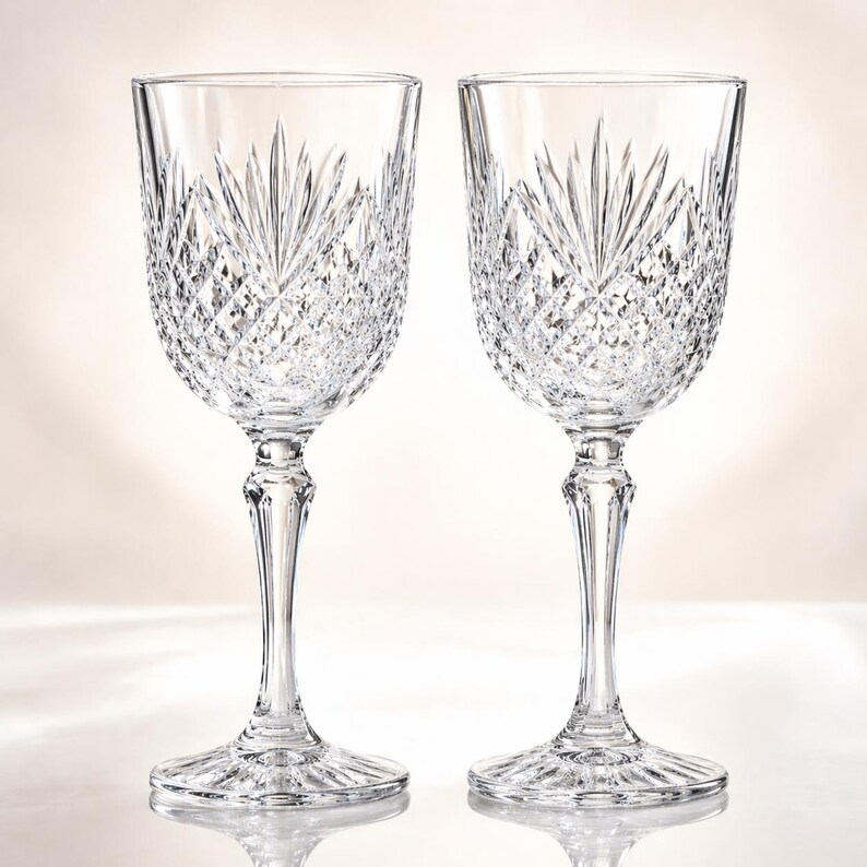 May include: Two clear crystal wine glasses. Each glass features a detailed, cut-glass pattern on the bowl and stem. The bowl has a fan-like design at the top, with diamond-shaped cuts throughout. The stems are slender and elegant.