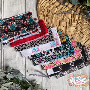 May include: A collection of colorful headbands with various patterns, including leopard print, floral designs, and text that reads "Like Mom" and "Mamas Girl". The headbands are arranged on a wooden surface with a woven basket in the background.