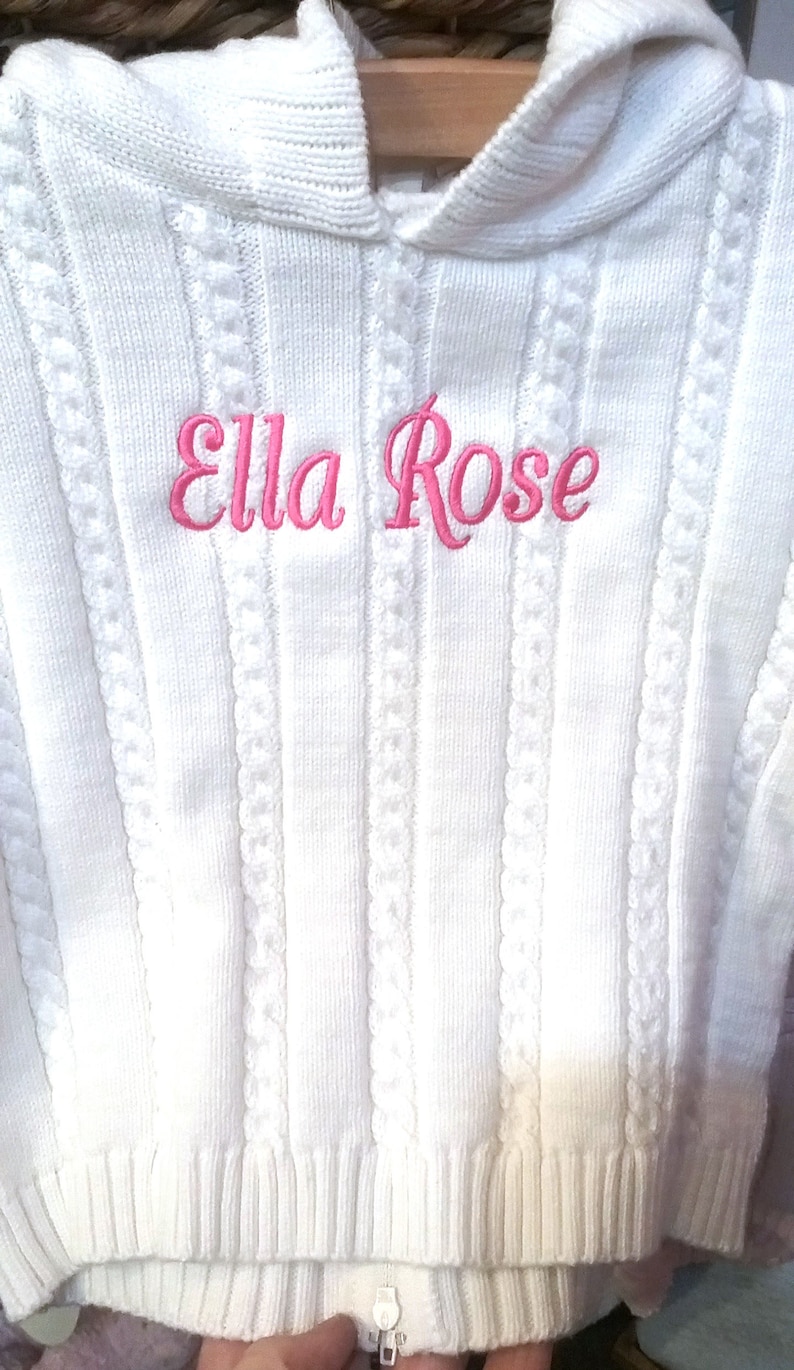May include: White knitted hooded sweater with a pink embroidered name "Ella Rose"