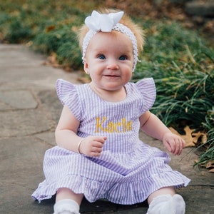 Puede incluir: Una niña pequeña con una diadema blanca con un lazo, un vestido de cuadros vichy morado y blanco con el nombre "Kacie" bordado en la parte delantera y calcetines blancos. Está sentada en un camino de piedra.
