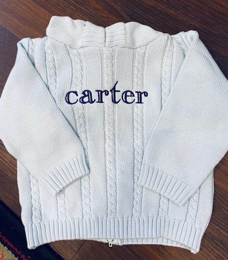 May include: A light blue knitted sweater with a zipper closure and a cable knit pattern. The sweater has the name "carter" embroidered on the front in dark blue thread.