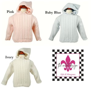 May include: Three hooded cable knit sweaters in pink, baby blue, and ivory. The sweaters have a ruffled trim around the hood. The sweaters are displayed on mannequins. The image includes the text "the initial design Gifts & Monograms".