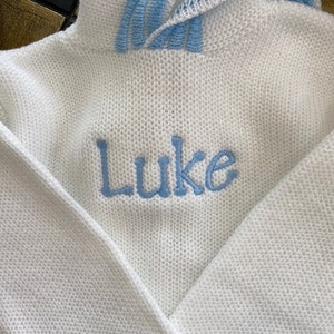 May include: White knitted sweater with blue trim and the name "Luke" embroidered on the front.