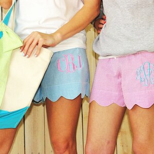 May include: Two women wearing matching blue and pink striped shorts with scalloped edges. The shorts have embroidered monograms. One woman is holding a white tote bag with a green and blue bow.