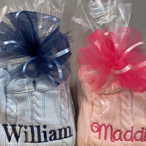 May include: Two personalized cable knit sweaters, one light blue with the name "William" embroidered in dark blue, and one pink with the name "Maddi" embroidered in pink. Both sweaters are wrapped in clear plastic with a bow.