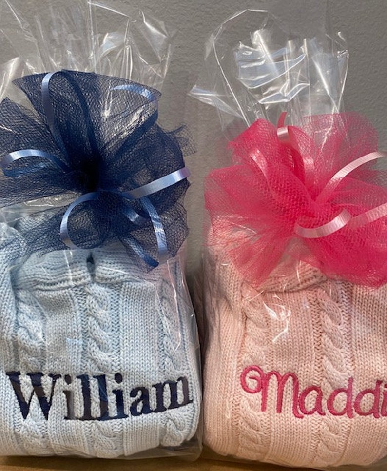 May include: Two personalized cable knit sweaters, one light blue with the name "William" embroidered in dark blue, and one pink with the name "Maddi" embroidered in pink. Both sweaters are wrapped in clear plastic with a bow.