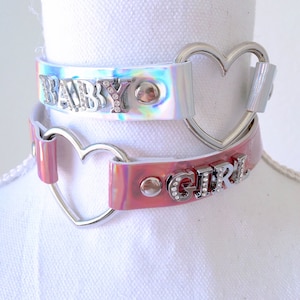 May include: Two iridescent choker necklaces with heart-shaped clasps. The top choker is silver with the word "BABY" in rhinestones. The bottom choker is pink with the word "GIRL" in rhinestones.