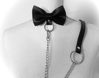 May include: A black leather bow tie with a silver chain and a matching leather strap. The bow tie is attached to the chain with a silver ring.