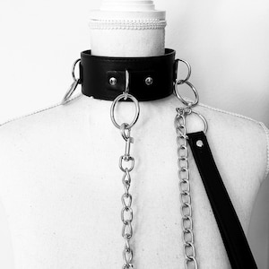 Puede incluir: Un collar de cuero negro con anillos de metal plateado y una correa de cadena. El collar es ancho y se asegura con tachuelas de metal. La cadena y la correa están unidas al collar con grandes anillos de metal. Se muestra en un maniquí blanco.