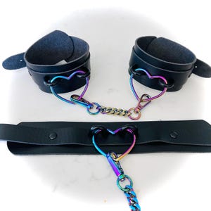 May include: A set of black leather cuffs with iridescent heart-shaped metal accents connected by a chain. The cuffs are attached to a black leather choker with a heart-shaped pendant.