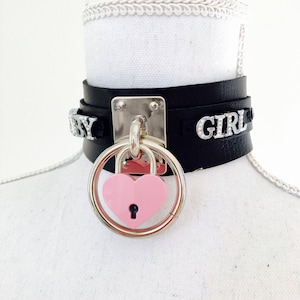 Puede incluir: Choker de cuero negro con un candado plateado en forma de corazón y las palabras "Baby Girl" en pedrería plateada.