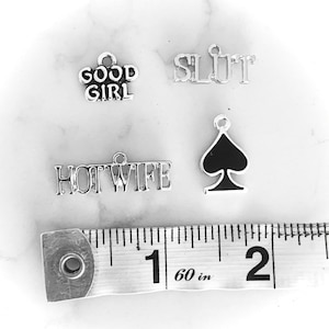 May include: Four silver charms with different words and a black spade charm. The charms are on a white surface with a measuring tape that reads 60 in.