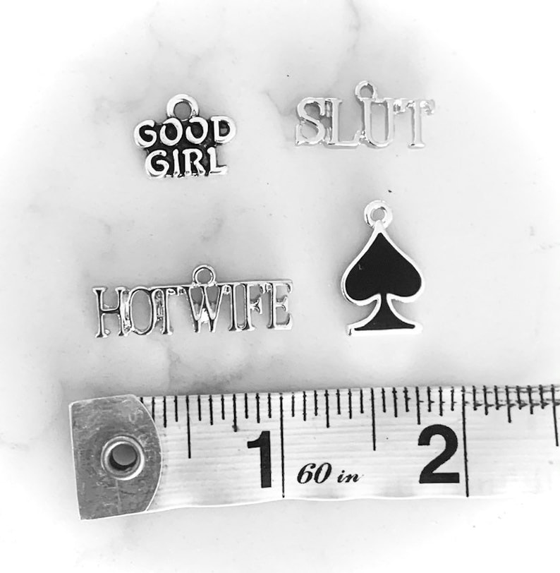May include: Four silver charms with different words and a black spade charm. The charms are on a white surface with a measuring tape that reads 60 in.
