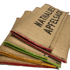 May include: Stack of four burlap bags with colorful trim. The top bag has the text "NATHALIES APFEL SACK" printed on it.