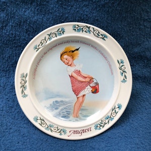 May include: A white ceramic plate with a blue floral border. The center of the plate features a painting of a girl in a pink dress holding a bucket. The text "A certified plate in the original and only Sarah Stilwell Weber Calendar Collection" and "August" are printed on the plate.