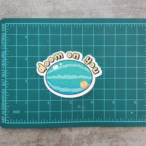 May include: A sticker featuring a cartoon watermelon with the words "doom on you" in a playful font. The watermelon is blue and green with a gold seed. The sticker is on a green cutting mat.