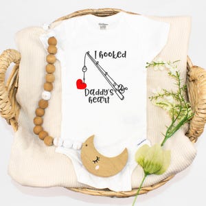 Puede incluir: Un body blanco para bebé con una ilustración en blanco y negro de una caña de pescar con un corazón rojo en el anzuelo. El texto "I hooked Daddy's heart" está impreso en el body.