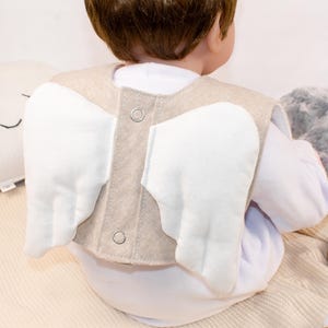 Puede incluir: Un chaleco beige de fieltro para bebé con alas de ángel de fieltro blanco unidas. El chaleco tiene dos anillos metálicos en la espalda.