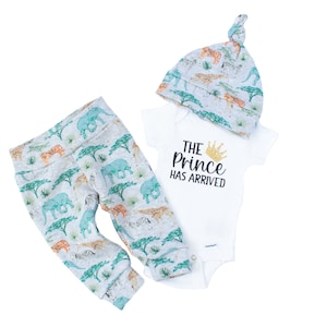 May include: A baby outfit set with a white bodysuit, grey and green patterned trousers, and a grey and green patterned hat. The bodysuit has the text "THE PRINCE HAS ARRIVED" with a gold crown.