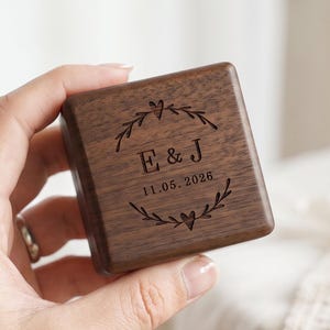 May include: A square dark brown wooden box with a smooth finish. The lid features a laser-engraved design of a circular wreath with two small hearts, the letters E & J, and the date 11.05.2026 centered within the foliage pattern.