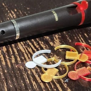 Può includere: Un flauto dolce nero Tudor con una fascia argentata e una clip rossa fissata sulla testata, accompagnato da un piccolo mucchio di clip circolari in plastica sciolte di colore bianco, oro e rosso sparse nelle vicinanze.