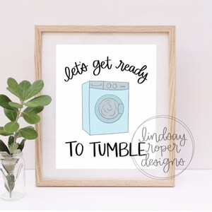May include: A white framed print with a blue and white cartoon illustration of a washing machine. The text "let's get ready to tumble" is written in black cursive.