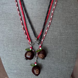 May include: A layered necklace set featuring three dark brown horse chestnuts as pendants. The cords are made of black, red, and white braided rope, accented with small green leaf-shaped beads and tiny white and red spherical beads.